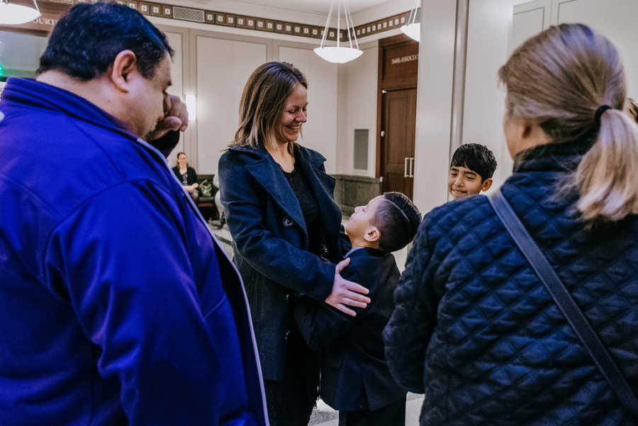 Adoptive mother holds shoulder of adopted son with people crying in background at adoption court
