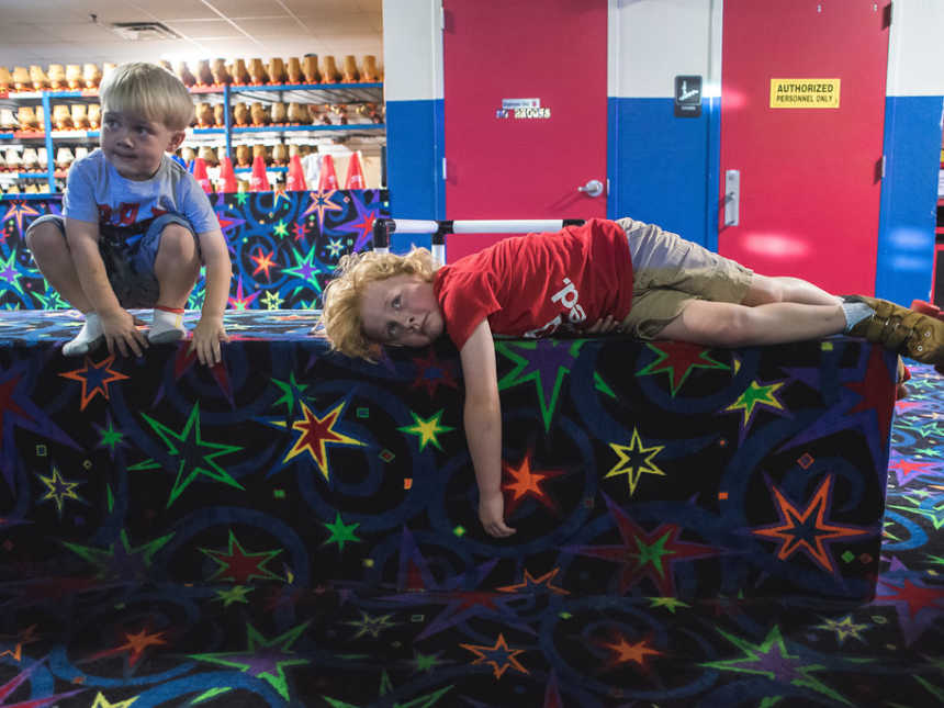 Young boy squatting on neon carpeted bench at roller rink while another lays on his side next to him