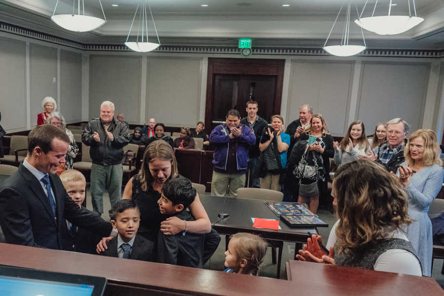 Mother and father hugging adopted sons with birth children and people in crowd watching at adoption court