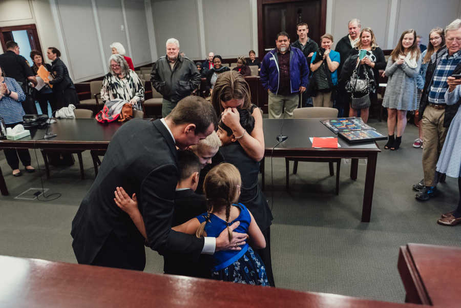 Adoptive mother and father hug adopted sons with two birth children at adoption court while people watch smiling