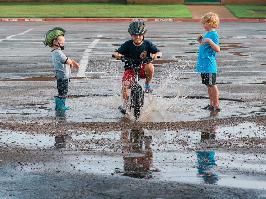 Boy riding his bike in a large puddle while two brothers stand in the puddle waiting to be splashed