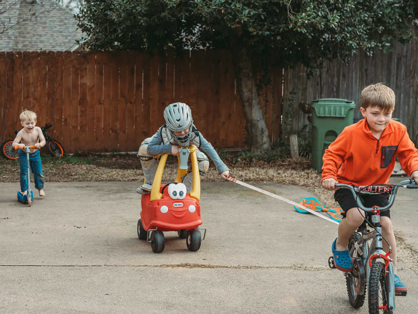 Brother riding bike pulls his brother who is sitting on toy car while youngest brother rides scooter shirtless in driveway