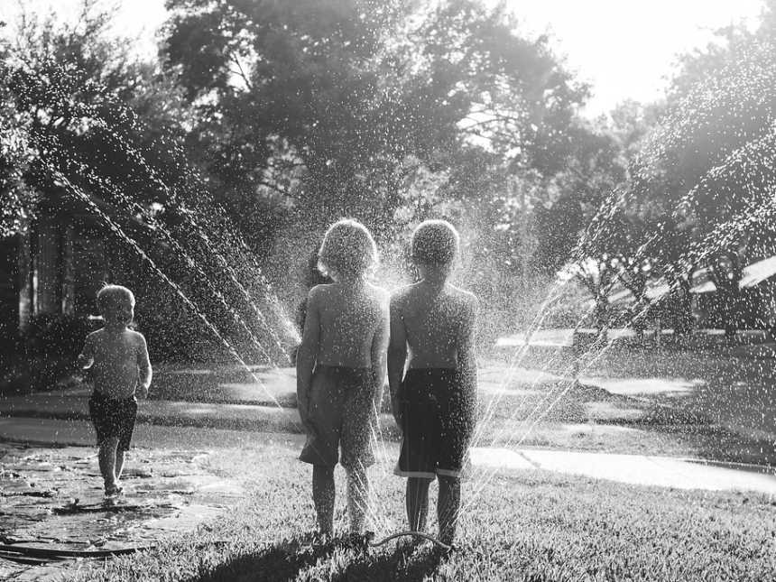 Two brothers stand side by side in yard in front of sprinkler while younger brother runs towards them