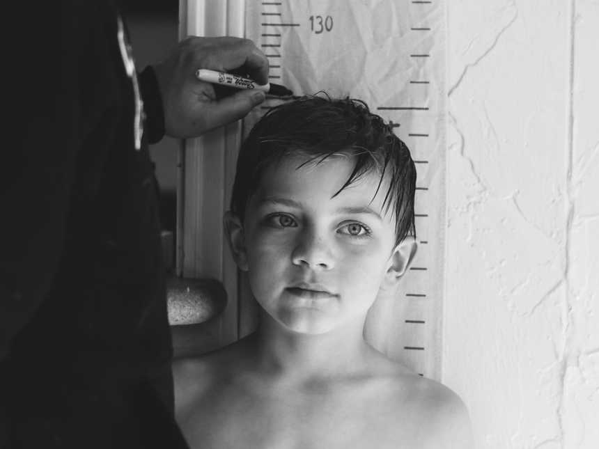 Boy standing against wall with measurements on it while older man makes a mark with sharpie above his head
