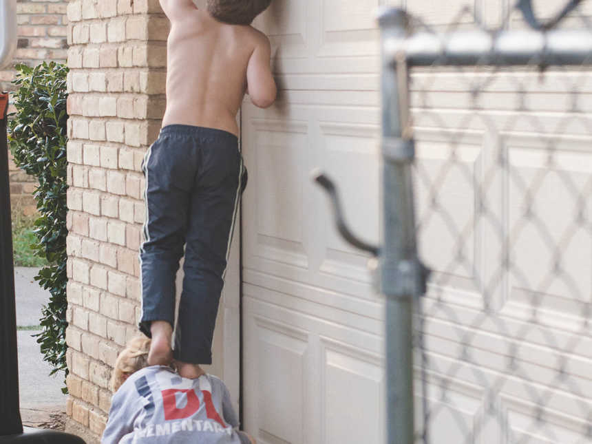 Little boy crouches on ground while brother stands shirtless on his back leaning on garage