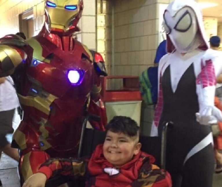 Boy with cancer smiles in wheelchair with iron man standing to his left and another comic book character to his right