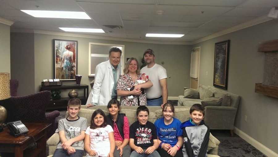 Husband and wife standing with newborn next to OBGYN with 6 adopted children sitting on couch in front of them