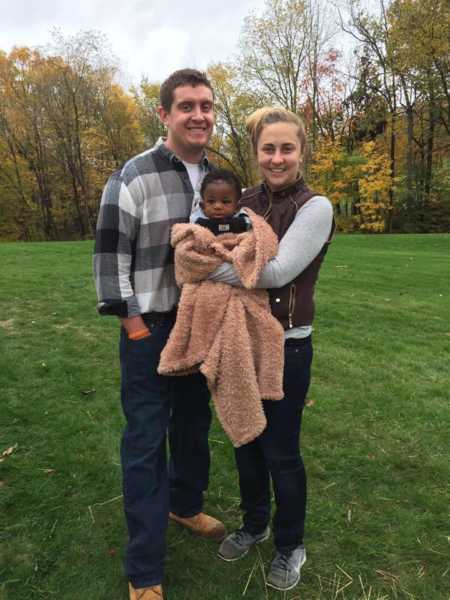 Husband and wife stand smiling in grass with their adopted son wrapped in blanket