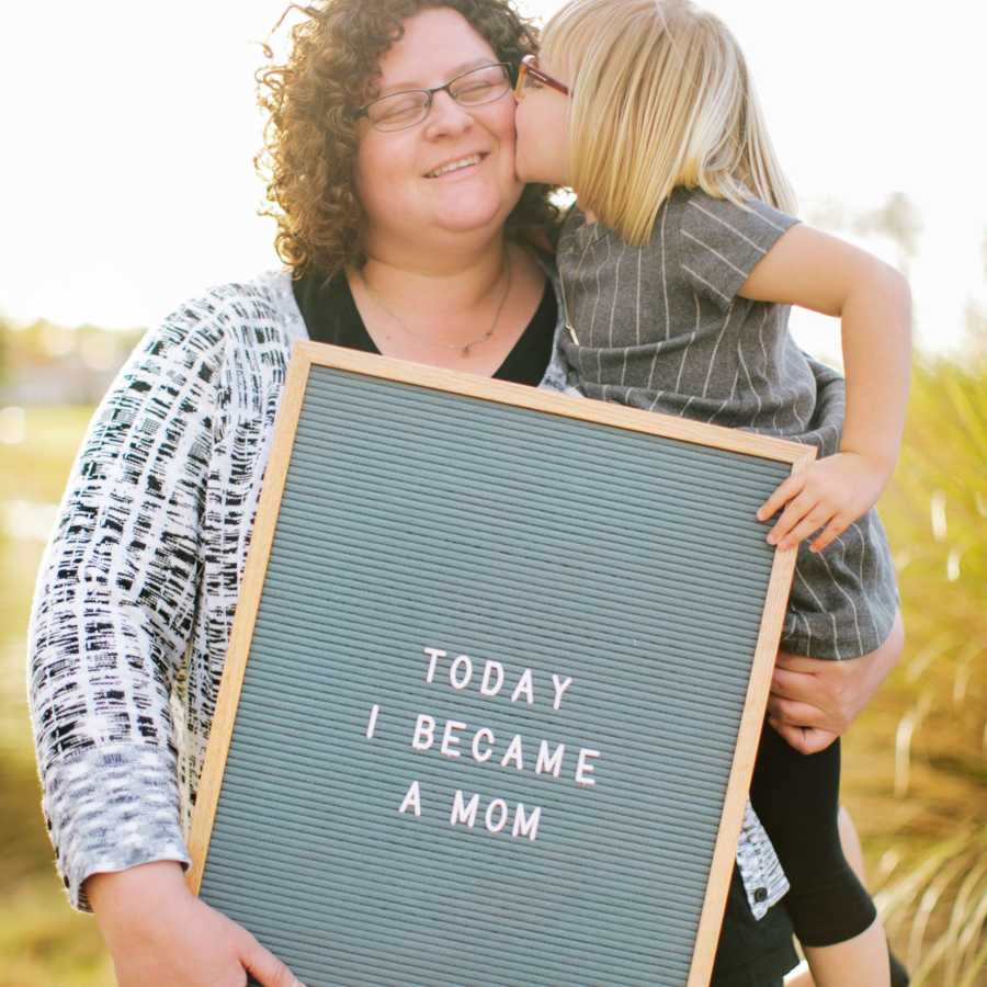 Mother holds adopted daughter and sign saying, "Today I became a mom"