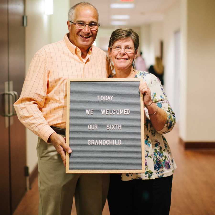 Grandparents of adopted child hold sign saying, "Today we welcomed our sixth grandchild"