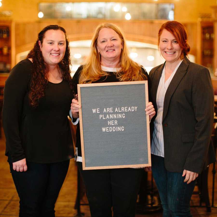 Three women standing in adoption court holding sign saying, "we are already planning her wedding"