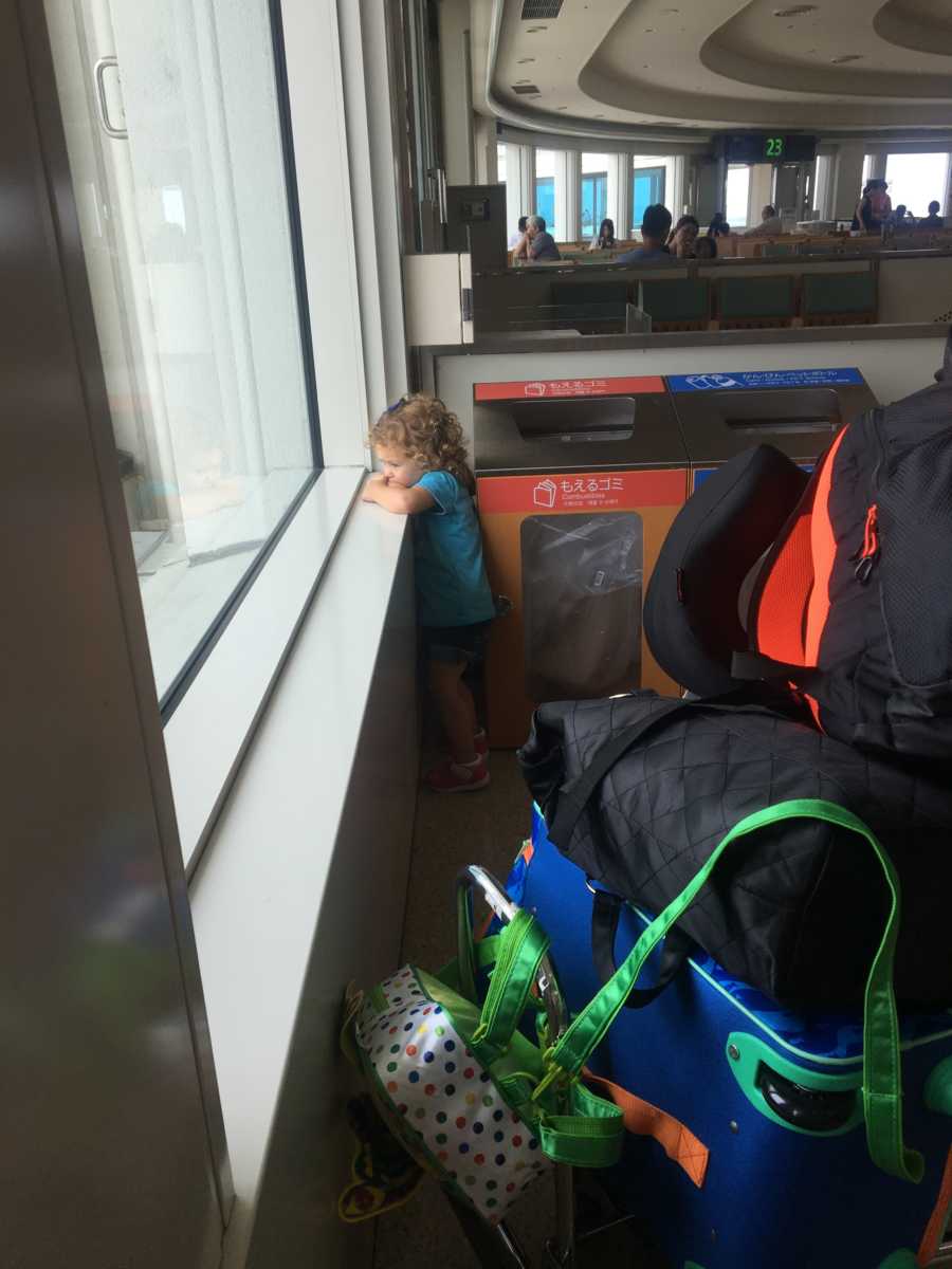 Little girl looking out window of airport with luggage behind her