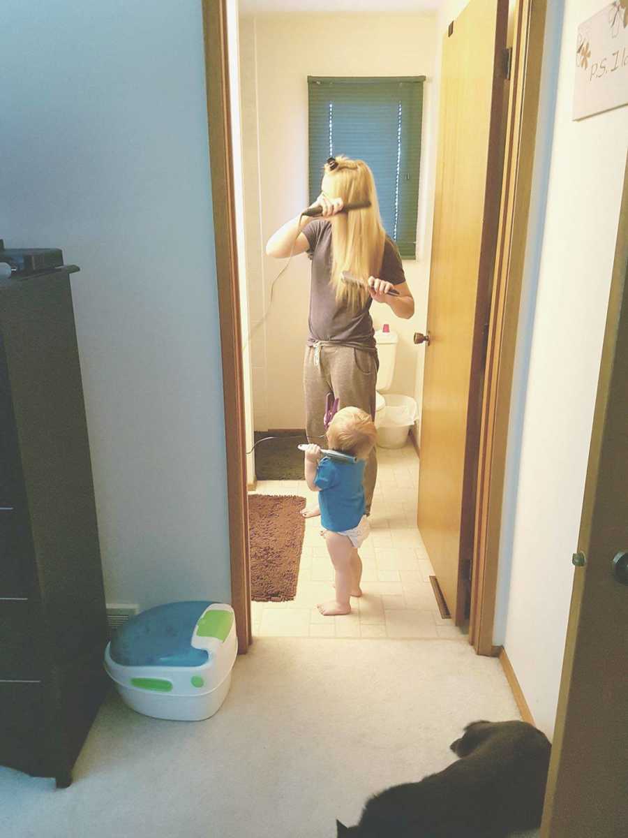 Mother straightening her hair in bathroom with toddler standing next to her mimicking her movements
