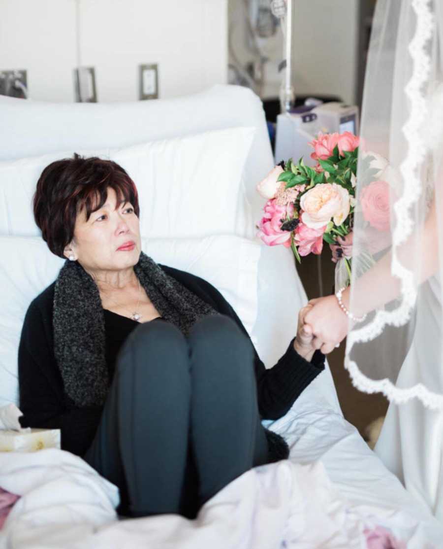 Terminally ill mother sits in hospital bed holding daughters hand who is in wedding gown 