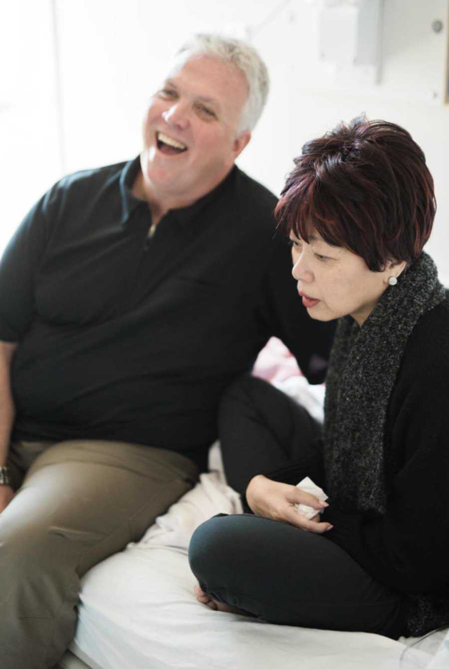 Terminally ill woman sits on hospital bed next to husband who is laughing