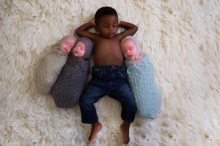 Adopted son sleeping with arms around his head with triplets on either side of him swaddled in blanket