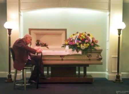Elderly husband sits besides deceased wife's casket