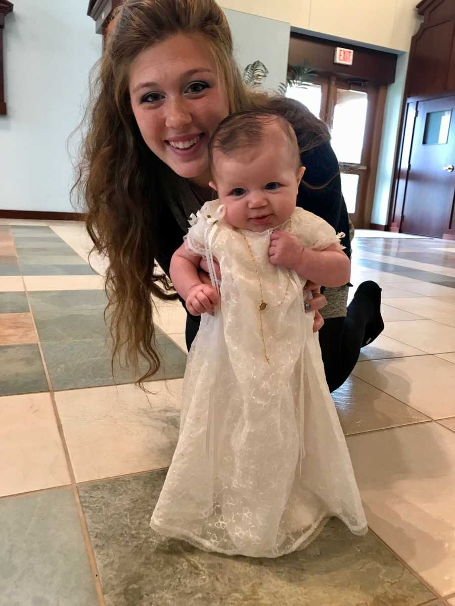 Mother sitting on ground holding up newborn who is wearing christening gown sewed by her grandmother