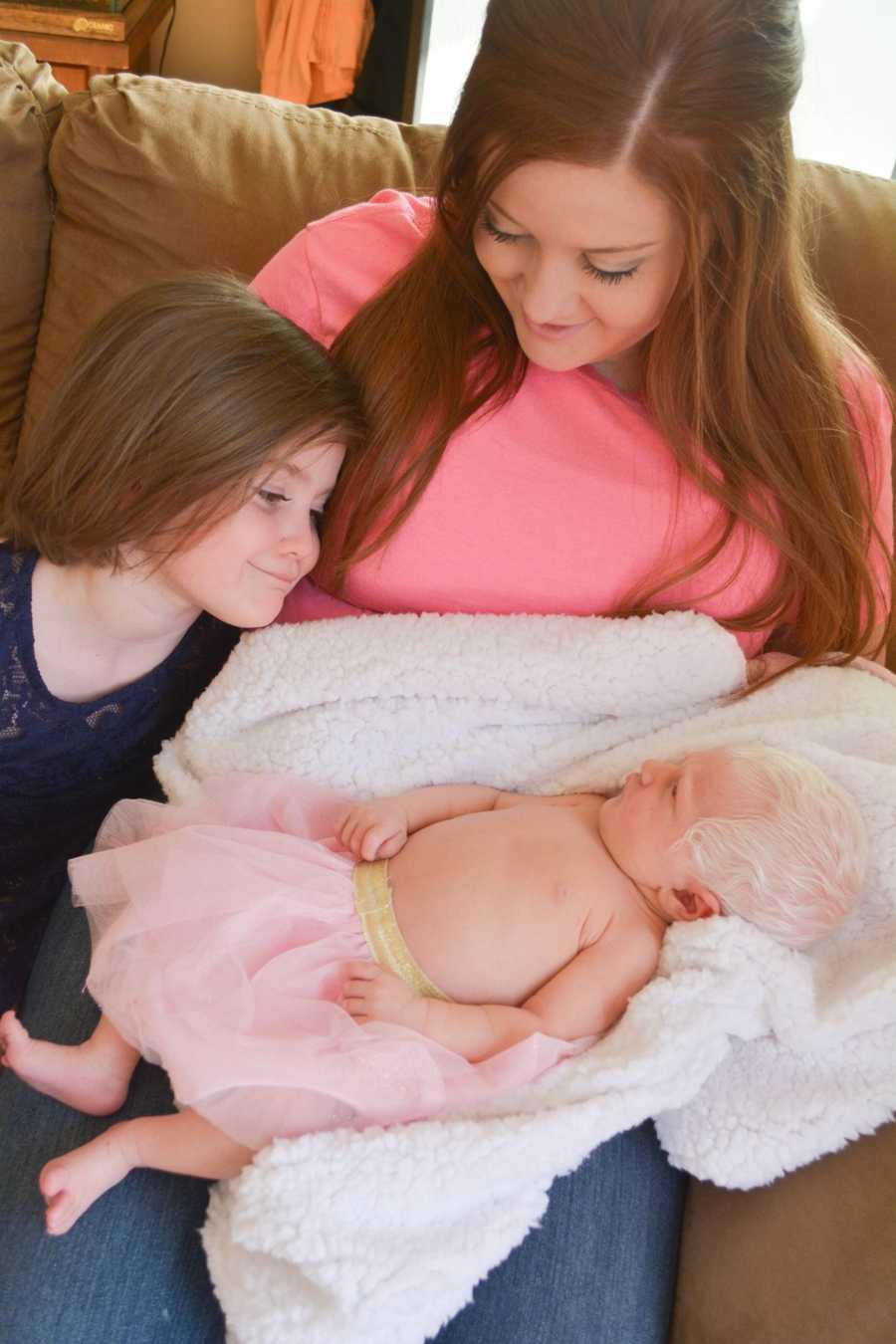 Mother holding albino newborn in lap with white hair in pink tutu while first born looks over at her
