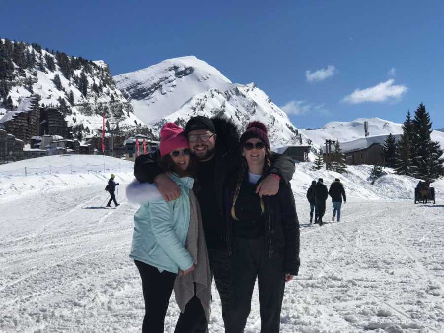 Two girls with their boy roommate smile on snowy slopes with mountains in background