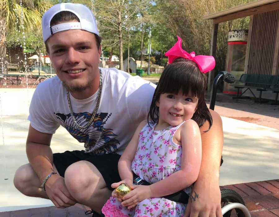 Man squats down smiling with arm around little girl in wheelchair 