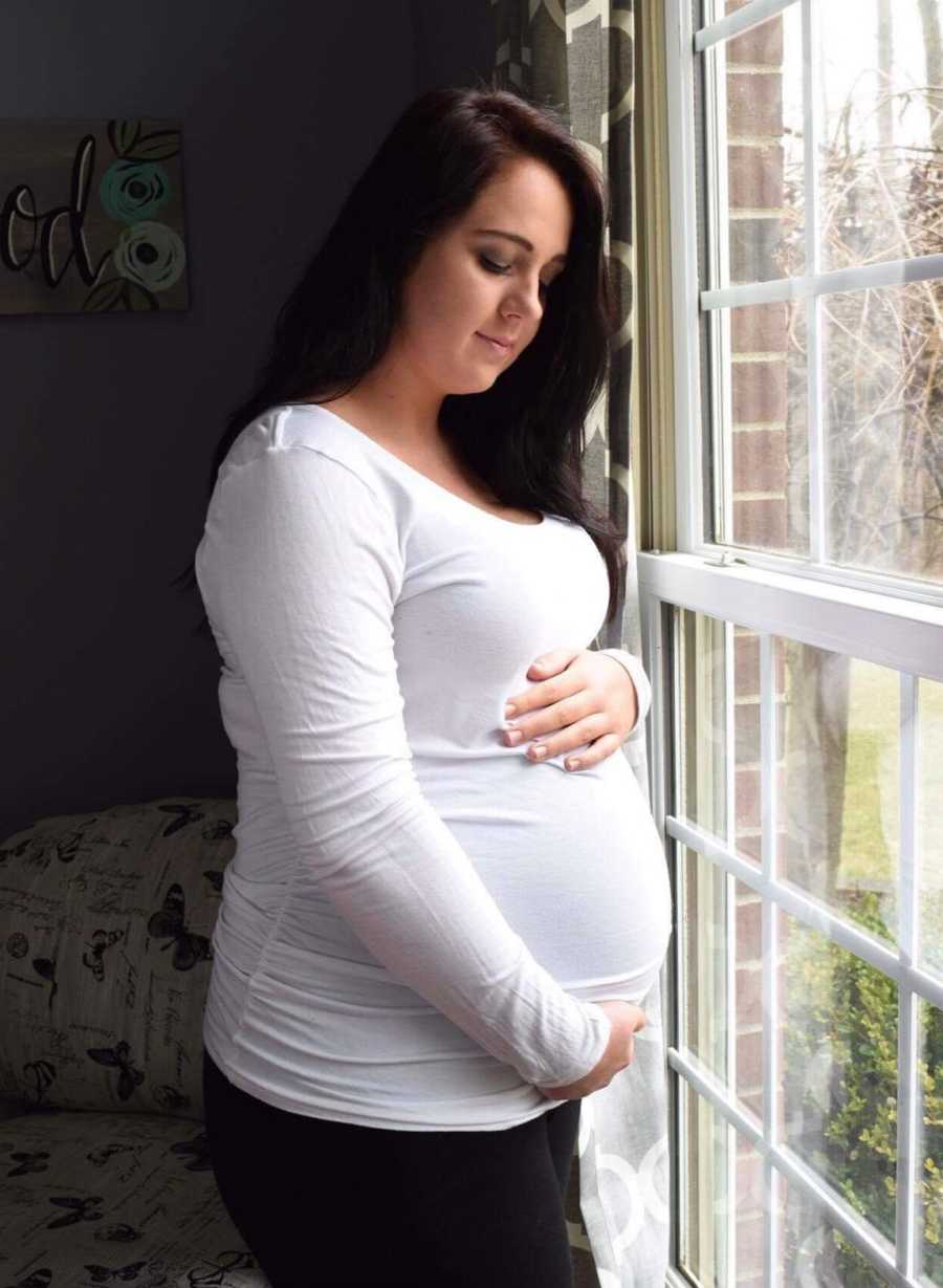 Pregnant woman holds stomach as she stands next to window in home