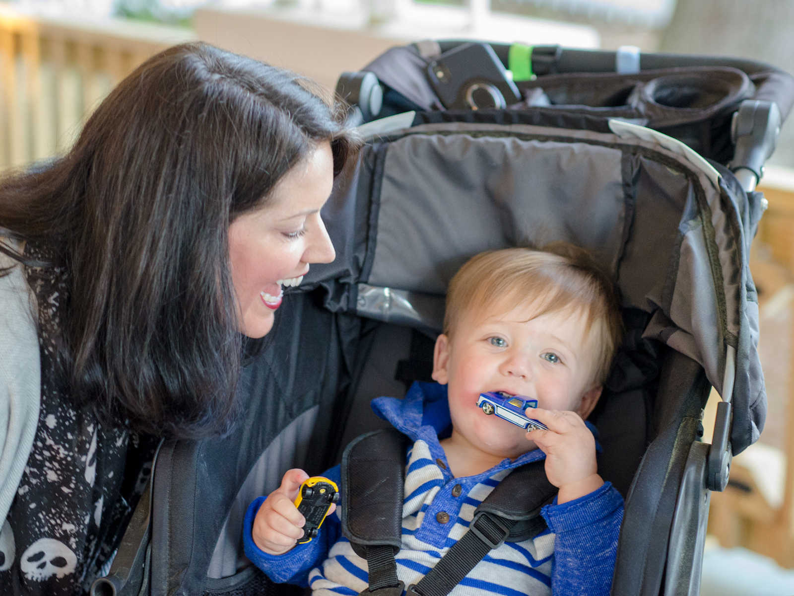 Mother smiles at adopted son sitting in stroller with toy car in his mouth