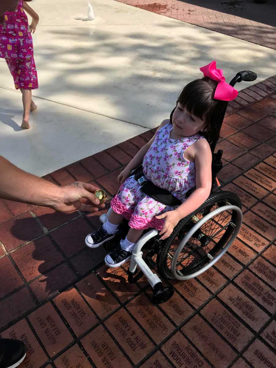 Man hands golden egg to toddler in wheelchair wearing large pink bow on her head