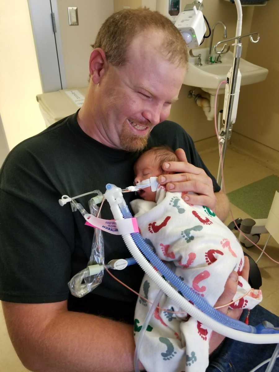 Father smiles while holding newborn close to chest in NICU with tubes attached to her face