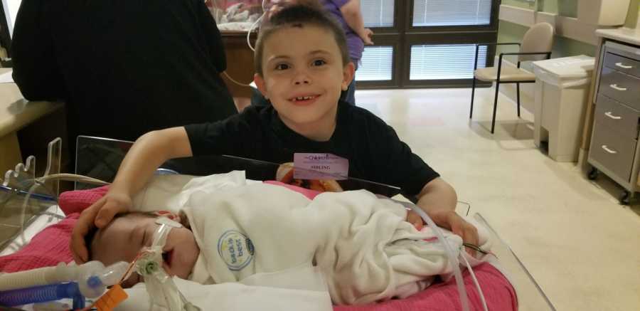 Toddler brother stands next to sister in NICU smiling