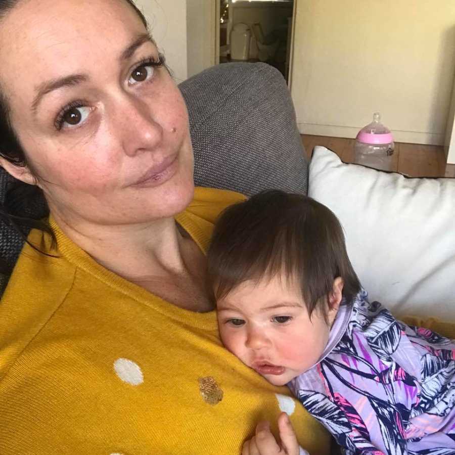 Mother who struggles to do it all sits on couch with infant daughter laying on her chest in selfie