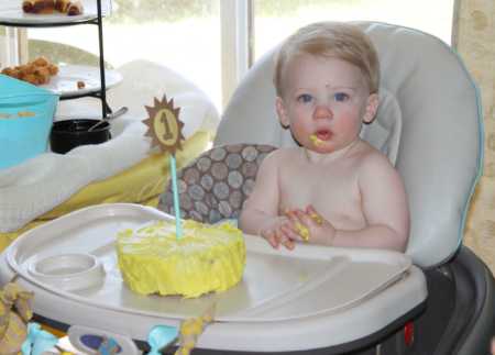 1 year old sitting in high chair with yellow birthday cake in front of her 