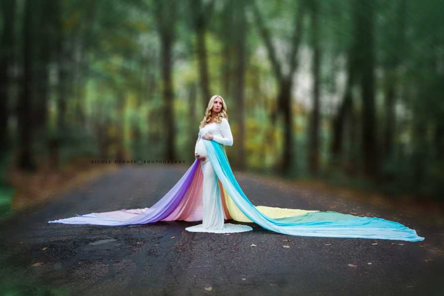 Pregnant woman stands in white dress with long rainbow train holding her stomach