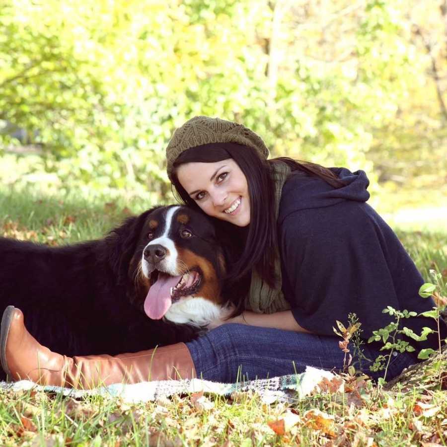 Woman sits in grass beside her Bernese mountain dog smiling