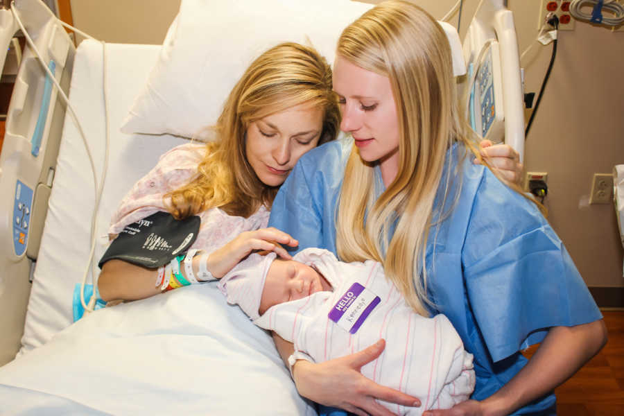 Wife holds newborn in her arms in hospital bed next to her wife who gave birth through IVF