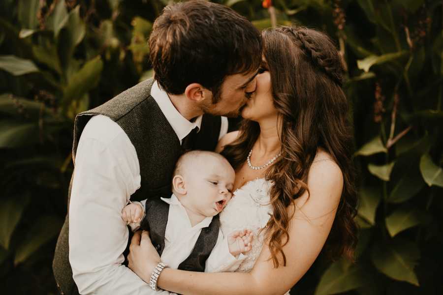 Bride and groom kiss while groom holds son who wants to breast feed