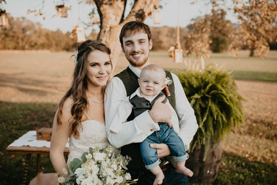 Bride and groom sit on picnic table with baby son in field