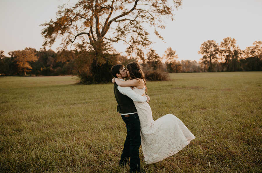 Groom holding bride in air while kissing her in grass field