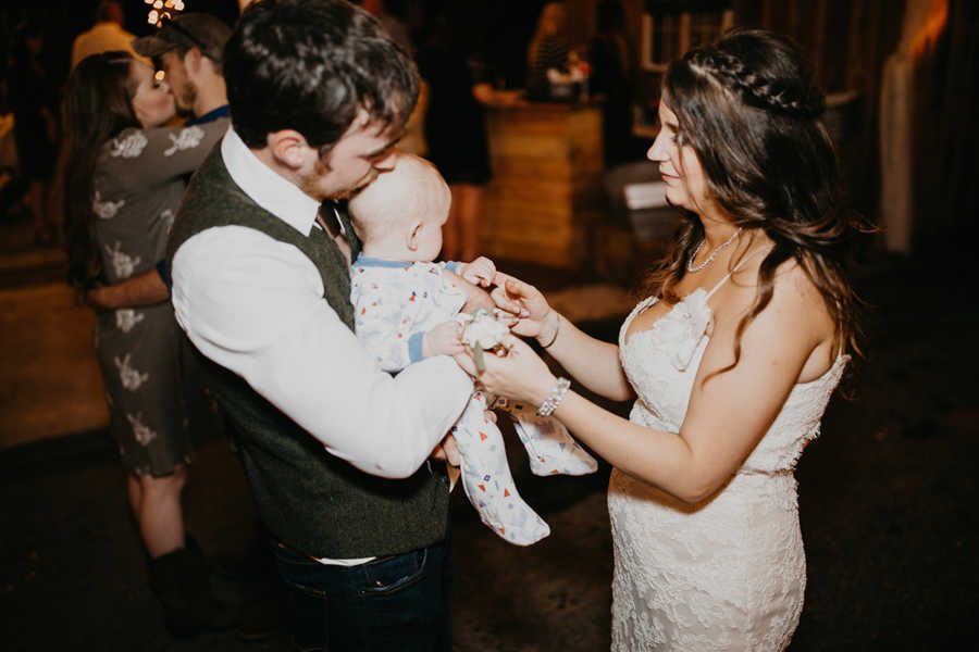 Groom holds son in onesie at wedding reception while bride holds sons hands
