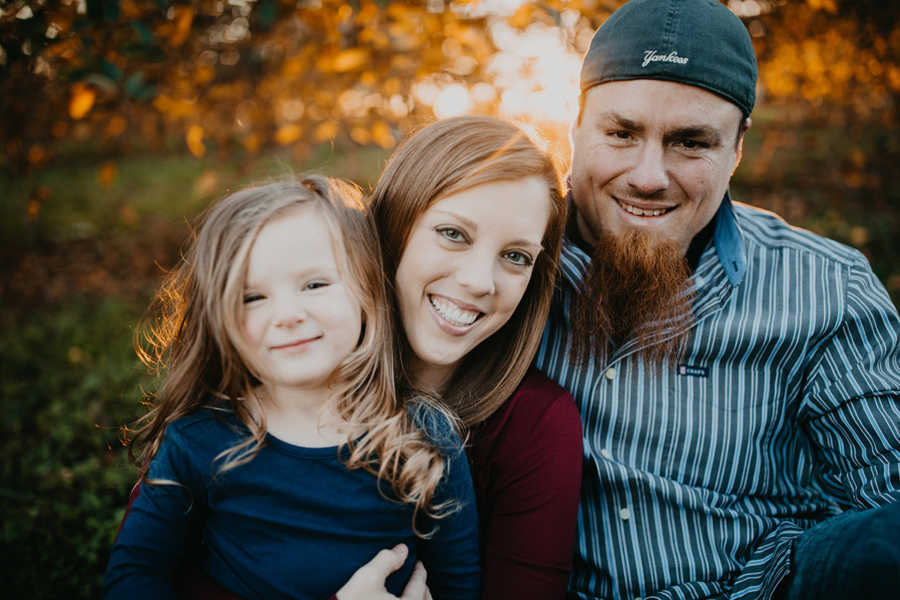 Mother with terminal breast cancer sits smiling with daughter in lap and husband to her right