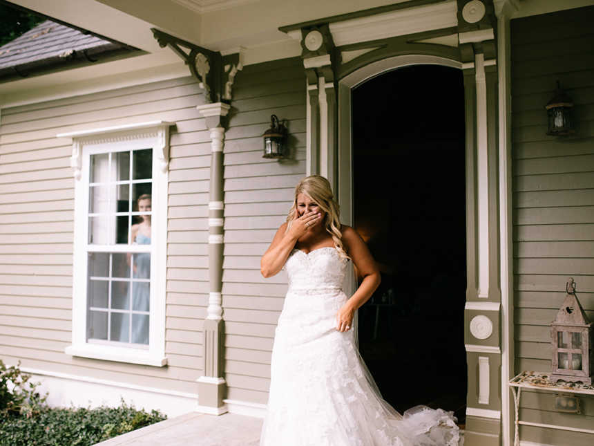 Bride holds hand to mouth crying as she walks out front door of house