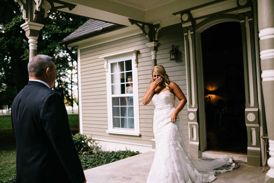 Bride holds hand to mouth crying as she walks out front door of house with dad awaiting her