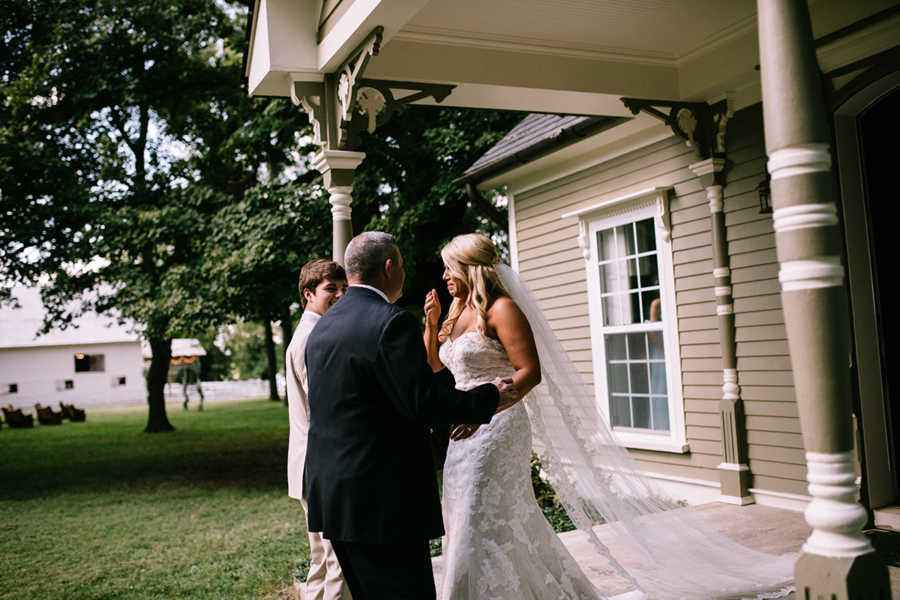 Bride stands on front porch crying as her dad reaches out for her with brother watching
