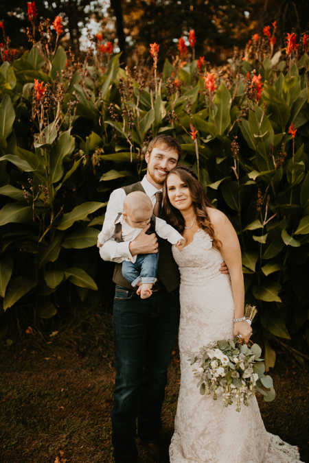 Bride and groom stand smiling in front of tall plants while groom holds baby son