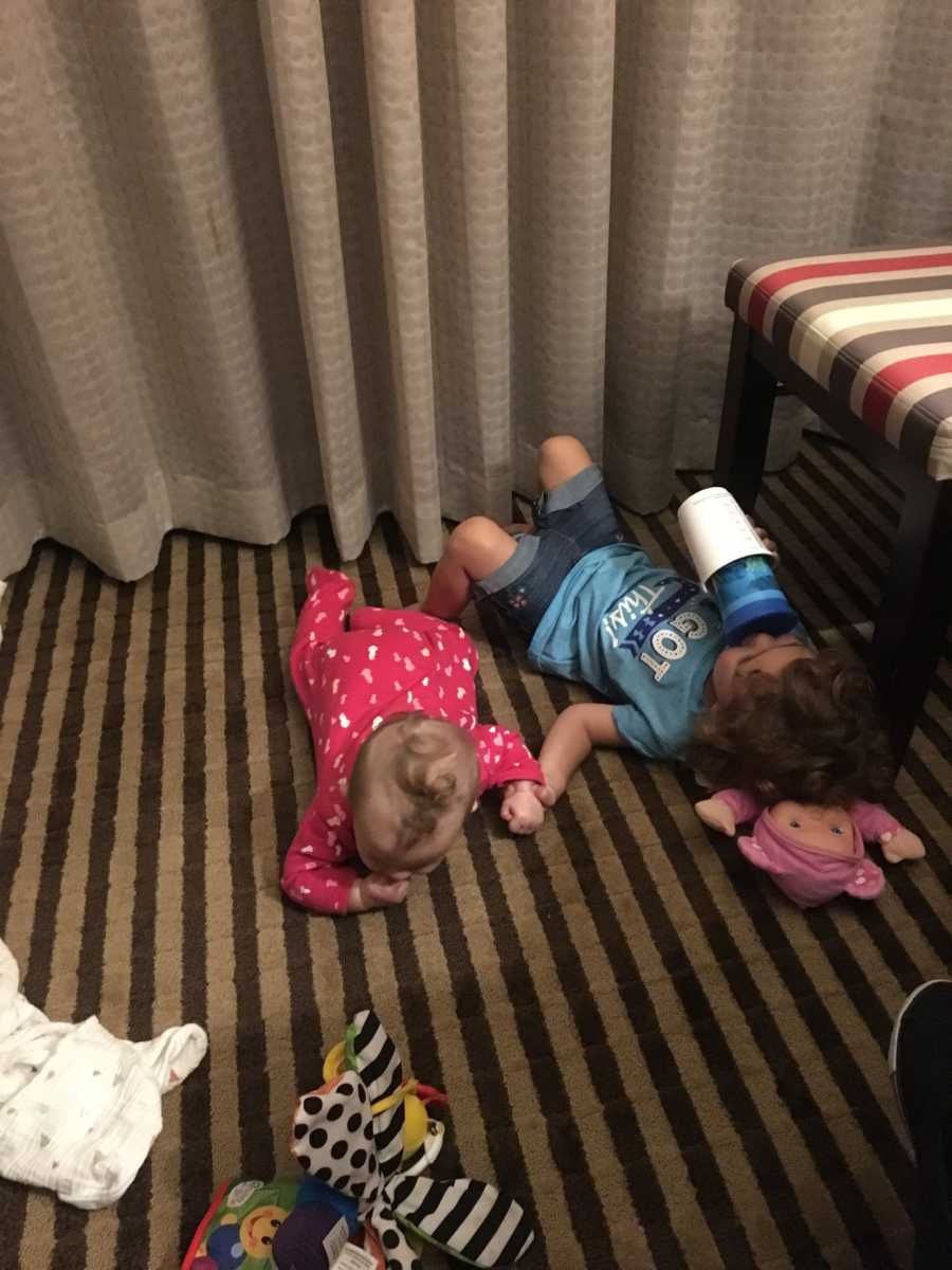 Toddler and infant siblings lying on floor of hotel room holding hands