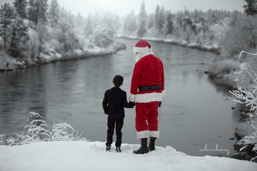 Boy stands in snow near body of water holding hands with mother's boyfriend in Santa suit