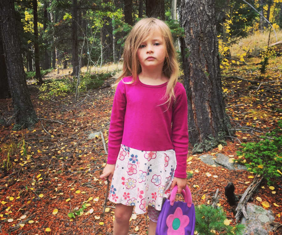 Toddler who mother says no problem saying no stands in forest with straight face holding little purse 
