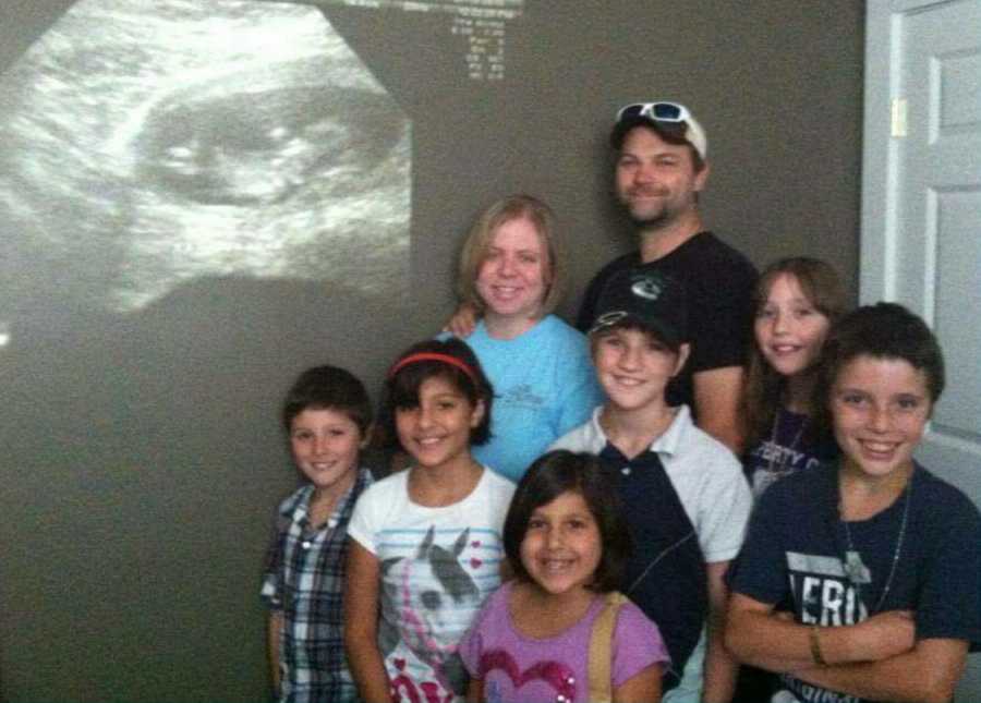 Husband and wife stand next to ultrasound projected on wall behind 6 adopted children