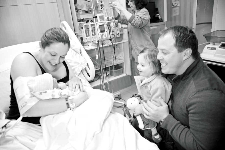 Woman who struggled with fertility holds newborn in hospital beside first born and husband