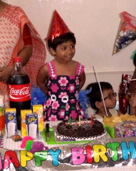 Adopted little girl wearing red party hat standing in front of cake at her birthday party